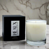 White candle in a glass container next to a black box with 'HEAUX' branding on a dark surface.